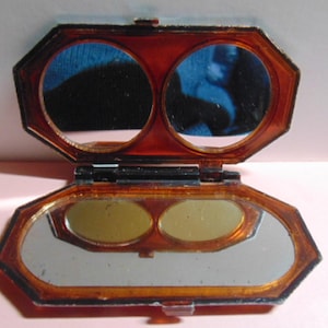 May include: Open, vintage, octagonal compact with a tortoiseshell-like frame. The compact has two round mirrors on top and two oval eyeshadow pans below. The bottom section has a large rectangular mirror.