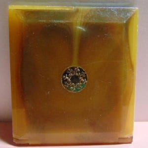 May include: A vintage, rectangular compact case in shades of amber and gold. The case has a decorative, circular gold emblem in the center. The compact appears to be made of a translucent material, with a slightly textured surface.