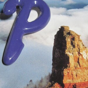 May include: A purple plastic letter P, shaped like a stylized letter, is placed on a background image of a rocky mountain landscape with clouds.