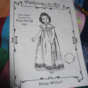 May include: A black and white illustration of a doll wearing a dress and a diaper. The illustration is on a white sheet of paper with the text "Fashions.to.Fit DOLL CLOTHES PATTERNS" at the top and "Betsy McCall" at the bottom.