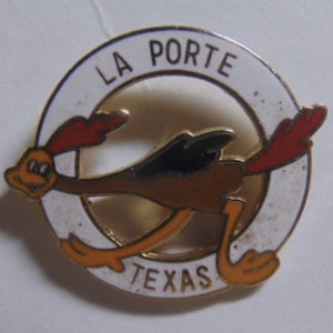 May include: A circular pin featuring a cartoon roadrunner in mid-sprint. The roadrunner is brown, orange, and black, with red accents. The pin has a white border with the words "LA PORTE TEXAS" in black lettering.