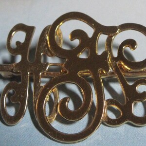 May include: Gold-toned metal brooch with a decorative design of intertwined letters, likely initials. The brooch has a pin back.
