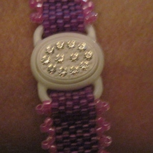 May include: A purple beaded bracelet with a white button closure featuring a circle of small rhinestones.