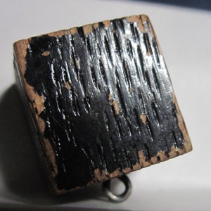 May include: A small wooden cube with a black painted surface. The paint is chipped and worn, revealing the wood underneath. The cube has a metal loop attached to it.