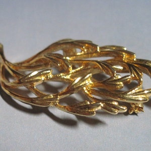 May include: A gold-toned brooch in the shape of a stylized leaf with intricate details. The brooch is made of metal and has a pin on the back for attaching to clothing.