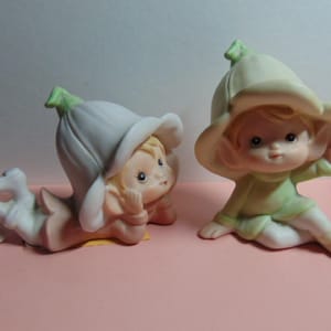 May include: Two ceramic figurines of flower fairies. One is lying down, the other is sitting. Both have blonde hair, rosy cheeks, and are wearing flower-shaped hats. The lying fairy is wearing a white outfit, and the sitting fairy is wearing a green outfit.