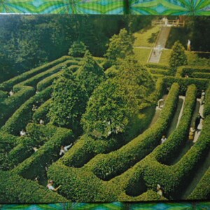 May include: An aerial view of a large green hedge maze with people walking through it. The maze is surrounded by trees and bushes.