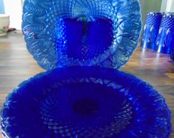 May include: A set of four cobalt blue glass dinner plates with a raised, textured design. The plates have a scalloped edge and a swirl pattern in the center.