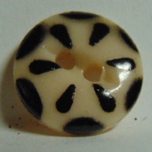 May include: A white button with a black starburst design. The button has a two-hole shank.