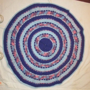 May include: A round crocheted rug with concentric circles in shades of blue, pink, and white. The rug has a center circle in a darker shade of blue.