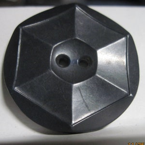 May include: A black, octagonal button with two holes in the center. The button has a slightly raised, diamond-shaped design.