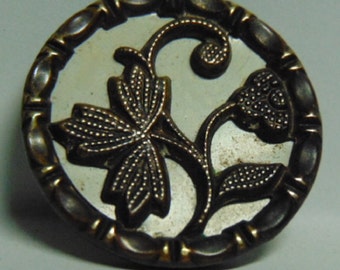 May include: A round, brass-colored button with a raised, ornate floral design. The button has a white center and a black floral pattern.