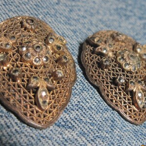 May include: Two antique, ornate, filigree metal brooches. Each brooch has a gold-toned metal frame with a detailed openwork design and is embellished with clear, sparkling rhinestones. The brooches are oval-shaped.