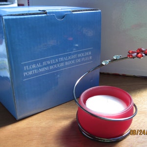 May include: A red glass tealight holder with a metal stem and a red jeweled flower. The holder is on a wooden surface and the box it came in is visible in the background. The box is blue and says "Floral Jewels Tealight Holder Porte-Mini Bougie Bijou de Fleur".