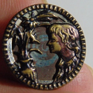May include: A vintage metal button with a gold-toned frame and a detailed image of a person with a plant behind them. The image is on a blue and green background.