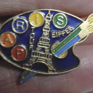 May include: A gold-toned metal pin shaped like an artist's palette with a blue enamel background. It features a gold Eiffel Tower, a paintbrush, and circular insets with the letters A, R, I, S, P, and S, along with the text TOUR EIFFEL.