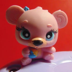 May include: A pink toy bear figurine with large blue eyes, long eyelashes, and a small blue bow. The bear is holding a small ball with yellow, blue, and orange sections. The figurine has large ears and a black nose.