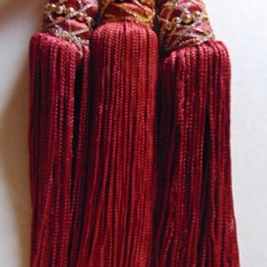 May include: Three decorative red tassels with gold accents. Each tassel features a bulbous top with gold beads and intricate gold threadwork. The long, flowing strands of the tassels are a deep red color, creating a luxurious look.