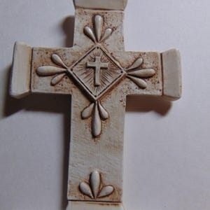 May include: Off-white cross with a detailed design. The cross features a central diamond shape with a smaller cross inside, surrounded by radiating lines. Decorative floral-like elements are placed around the diamond and on the arms of the cross.