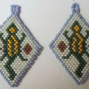 May include: Two square-shaped beaded ornaments with a floral design. The ornaments are white with green, yellow, and brown beads. The edges of the ornaments are light blue.