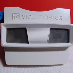 May include: A vintage View-Master viewer in white with a blue lever. The viewer has the words "View-Master" printed on the top. The viewer has two eye pieces and a center section with horizontal lines.