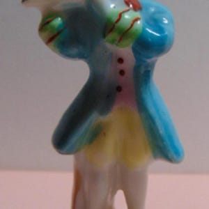 May include: A small porcelain figurine of a man in 18th-century attire. The figure wears a light blue coat, yellow breeches, and white stockings. The figurine is holding a tray and has a painted face.