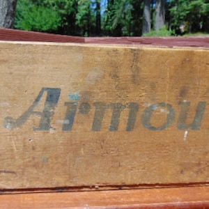 May include: A vintage wooden crate with the word "Armour" printed in black paint. The crate is made of rough-hewn wood and has a worn, rustic appearance.