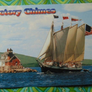 May include: A postcard featuring the schooner "Victory Chimes" sailing on blue water. The ship has large white sails and an American flag. A red and white lighthouse sits on a rocky island in the background. The sky is blue with some clouds.