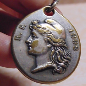 May include: A silver and gold oval pendant with a profile of a woman's face and the text "R.F. 1892".