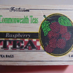May include: A wooden box of Commonwealth Teas Raspberry Tea. The box features the text "Raspberry TEA" in large letters, a graphic of raspberries, and the text "20 TEA BAGS" and "1.06 oz."