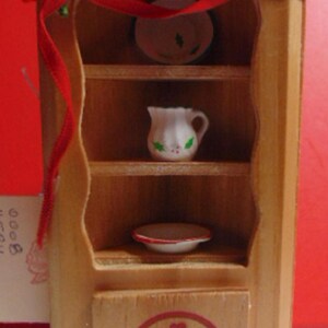 May include: A wooden miniature hutch with three shelves, a drawer, and a red bow. The hutch is decorated with a basket design and has a white pitcher and two white plates on the shelves.