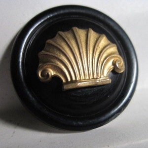 May include: A black circular button with a gold shell design. The shell is raised and has a detailed, ornate design.