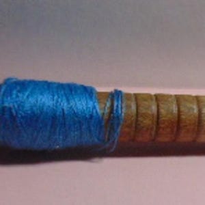 May include: A wooden spindle with three sections of colorful yarn. The yarn colors are orange, dark blue, and light blue. The spindle has a metal cap and is resting on a pink surface.