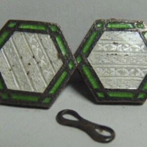May include: A pair of vintage cufflinks with a silver-colored hexagonal design, accented with green enamel and a dark border. The cufflinks have a textured pattern and are accompanied by a small, dark-colored connector.