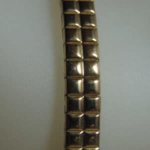 May include: Close-up of a gold-colored bracelet with a repeating square pattern. The bracelet is made of small, connected squares that create a flexible band. The surface of the bracelet has a shiny, reflective finish.
