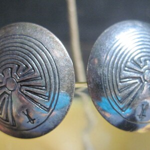 May include: A pair of silver earrings with a circular design featuring a maze-like pattern. The earrings are marked with an 'X' on the back.