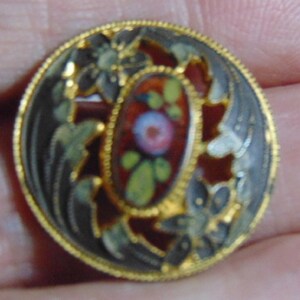 May include: A gold-toned button with a black and white floral design. The center of the button features a red, white, and green enamel flower.