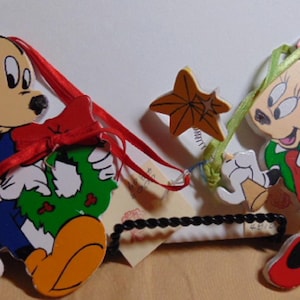 May include: Wooden Mickey Mouse and Minnie Mouse Christmas ornaments. Mickey wears a blue jacket and orange shoes, holding a green wreath with a red bow. Minnie wears a green dress and red shoes.