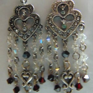 May include: Silver dangle earrings with a heart-shaped design, featuring clear and dark crystals. The earrings have multiple layers of chains and crystals.