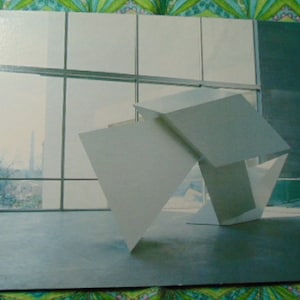May include: A white geometric sculpture made of three intersecting planes, photographed in a room with large windows. The sculpture is made of a smooth, white material and casts shadows on the floor.