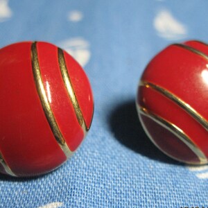 May include: Pair of red, round earrings with gold-colored stripes. The earrings have a glossy finish and are set against a blue background with a white pattern. These earrings are a classic accessory.