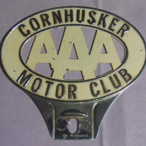 May include: A white and gold metal car bumper emblem with the text "Cornhusker AAA Motor Club" and the AAA logo. The emblem has a metal hook on the back for attaching to a car bumper.