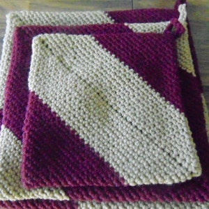 May include: Three knitted pot holders, one on top of the other. The top pot holder is beige with a burgundy diagonal stripe. The middle pot holder is burgundy with a beige diagonal stripe. The bottom pot holder is beige.