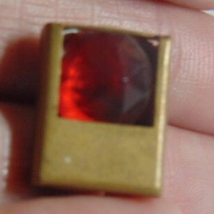 May include: A small, rectangular, brass-colored frame with a red glass insert.