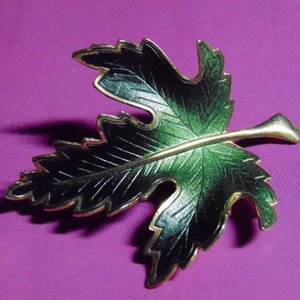 May include: A gold-toned leaf brooch with black and green enamel. The leaf has a detailed vein pattern and a pointed tip.