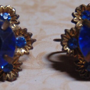 May include: A pair of gold tone earrings with a large blue rhinestone in the center of each earring. The rhinestones are surrounded by smaller blue rhinestones.