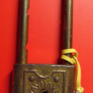 May include: A brass padlock with a keyhole and the text "SLAYMAKER" on the front. The padlock has a yellow rubber band attached to it.