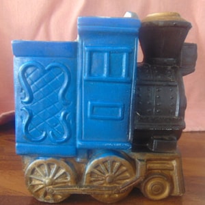 May include: A blue and black toy train with gold-colored wheels and accents. The train features decorative details on the side and a classic locomotive design. The toy train is a vintage item.
