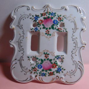 May include: White porcelain double light switch cover with floral design. The cover features a pink rose, blue and red flowers, and gold accents. The edges have a decorative, ornate design.
