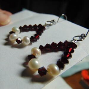 May include: A pair of dangle earrings with a red crystal and pearl design. The earrings are made with small red crystals and white pearls.